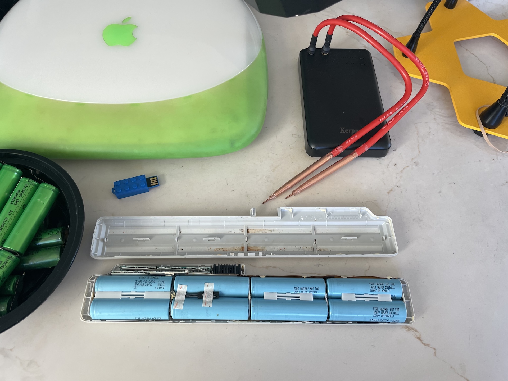 A workbench with a partially-completed battery, iBook, thumb drive, and spot welder