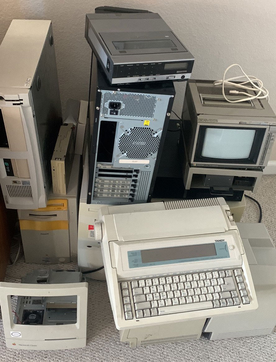 An empty Mac Classic case admist a pile of computers
